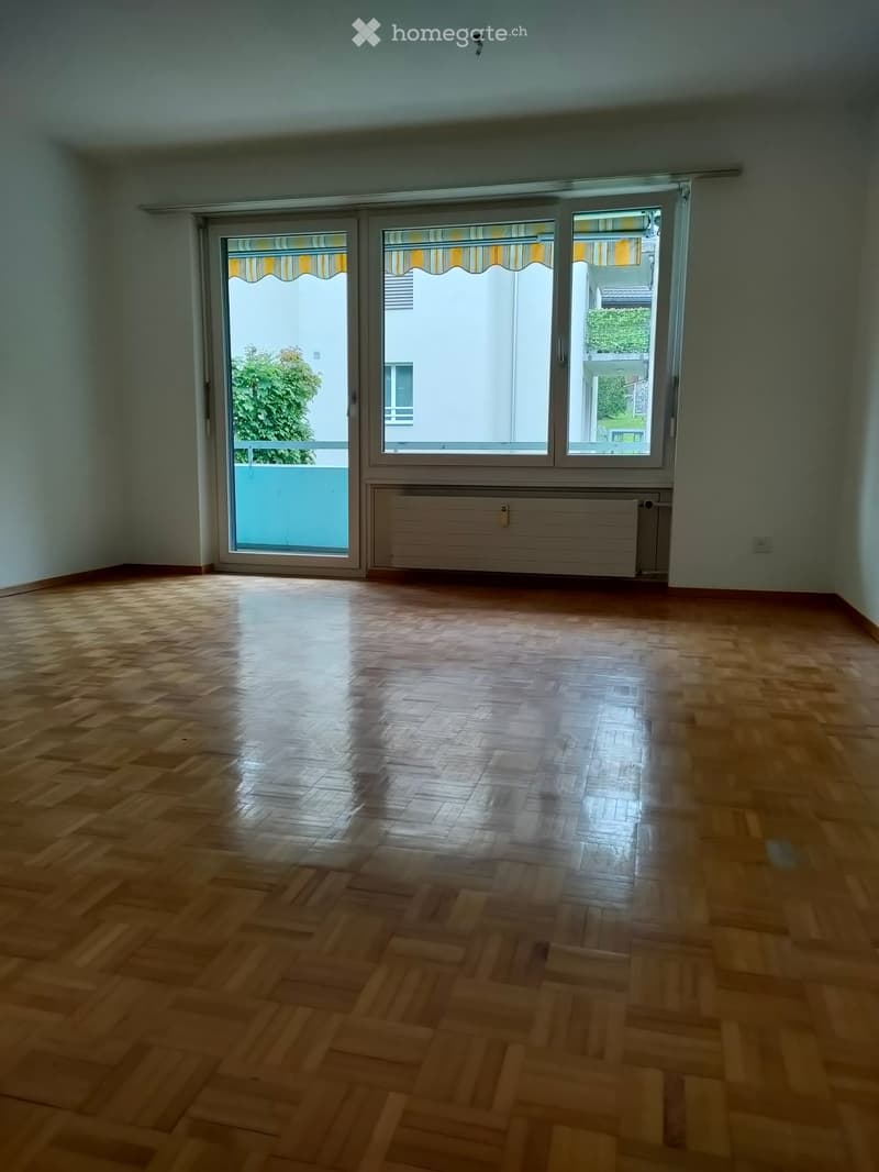 Empty room with wooden parquet flooring, large windows with blinds, radiator, and a view of greenery outside.