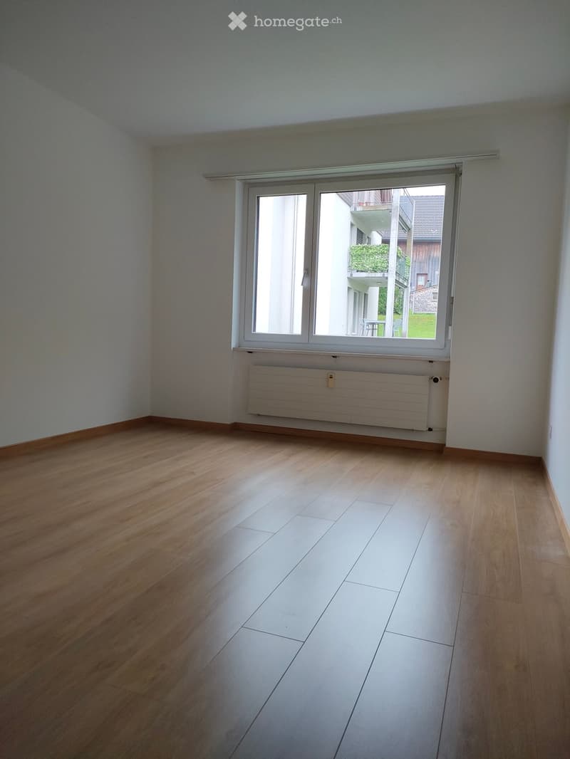 Empty room with wooden flooring, white tiled section, radiator, and large window with sliding doors leading to a balcony.
