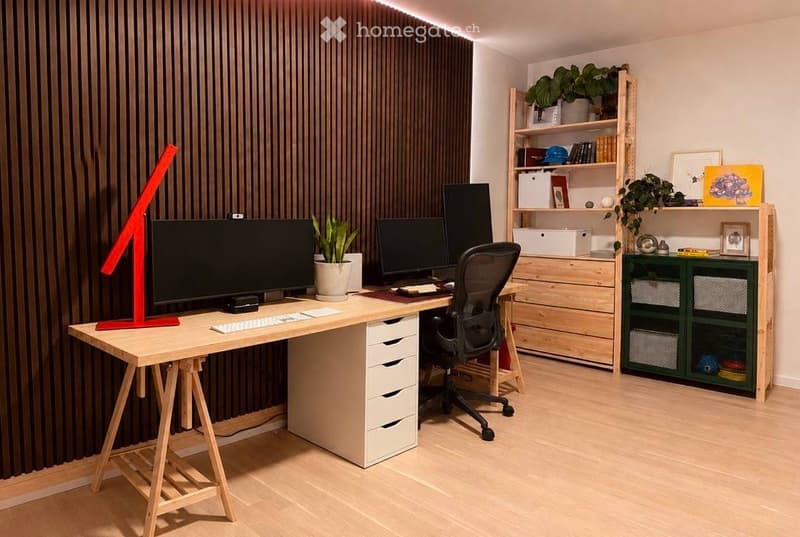 A modern, well-organized home office with a large wooden desk, multiple computer monitors, a comfortable office chair, and a variety of storage solutions including shelves and cabinets. The room features a sleek design with a combination of natural wood a