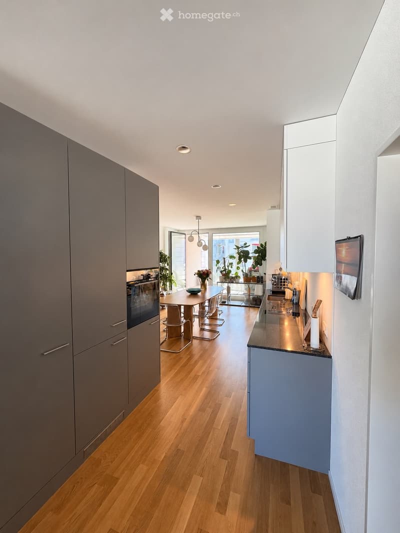 Modern kitchen with light wood flooring, grey and white cabinets, a central island with sink and faucet, a dining table with chairs, and decorative plants near the window.