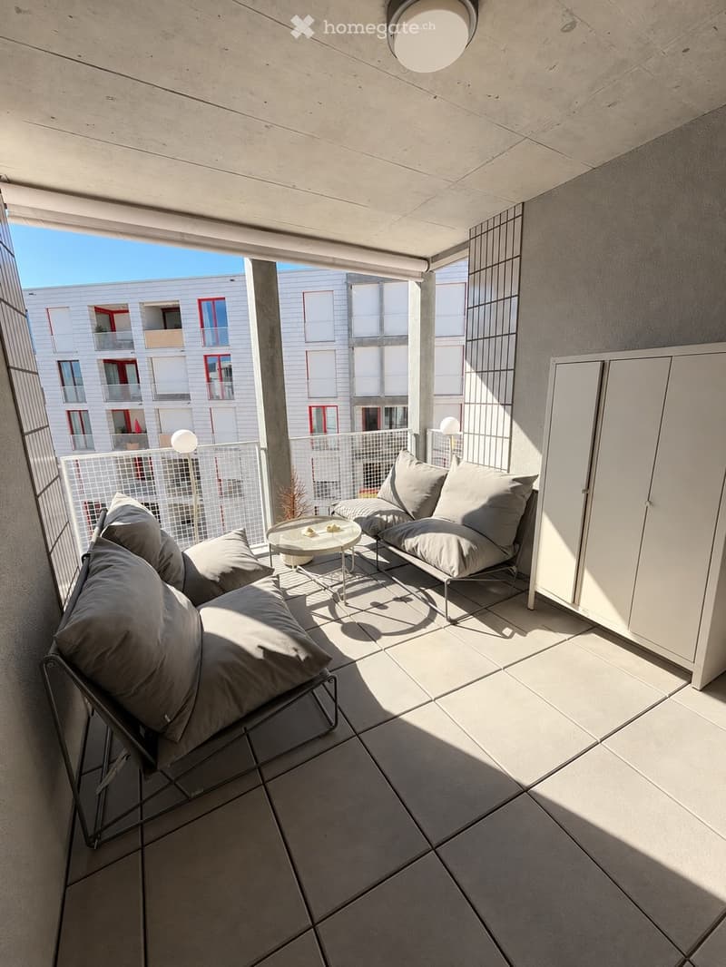 Modern balcony with tiled floor, two lounge chairs with grey cushions, small round table, and a view of neighboring buildings.