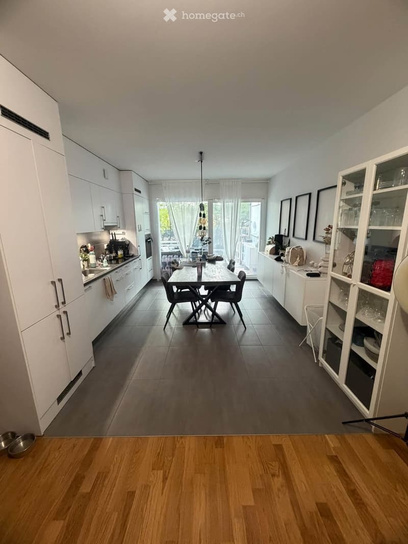 Modern kitchen with white cabinets, stainless steel appliances, a central dining table with chairs, and a large window with sheer curtains.