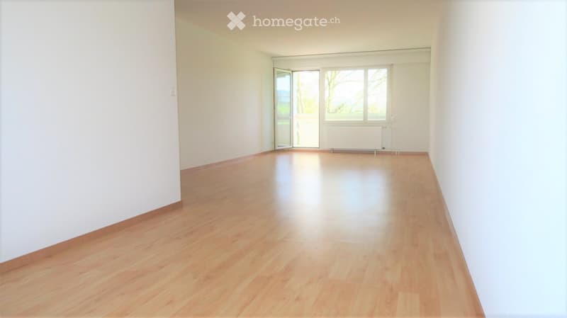 Empty room with light wooden flooring and white walls, featuring large windows and a sliding glass door leading to a balcony.