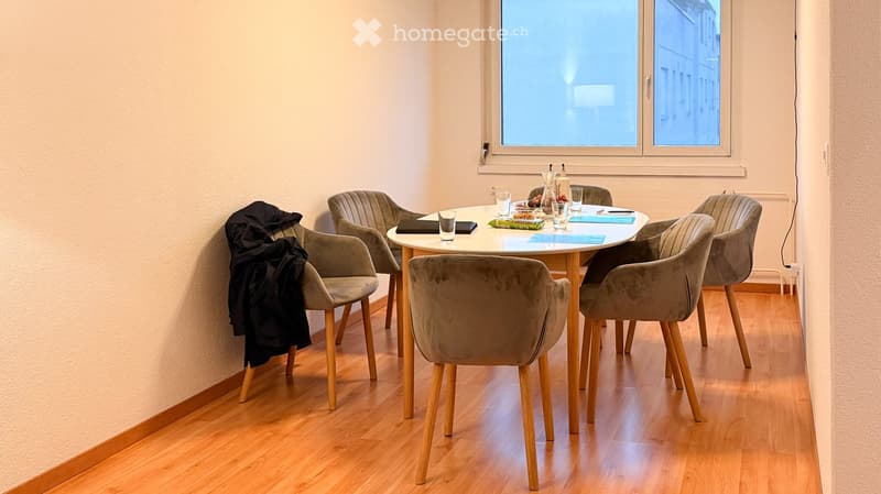 A modern dining area with a round wooden table surrounded by six upholstered chairs, set with glasses, a bottle, and placemats. The room features light wooden flooring and large windows.