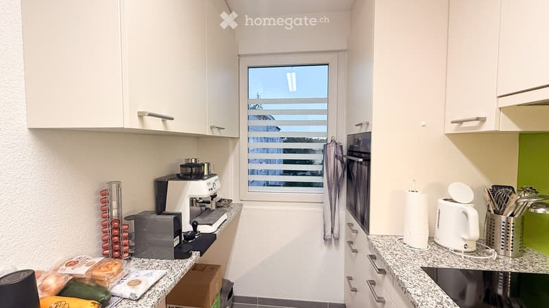 Modern kitchen with white cabinets, granite countertops, and various appliances including a coffee maker, toaster, oven, kettle, and knife holder. There is a window with blinds and a view of the outside.