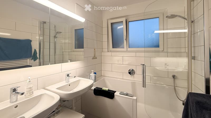 Modern bathroom with two sinks, a bathtub, a large mirror, and a glass-enclosed shower area. The room features white tiles and a window with frosted glass.