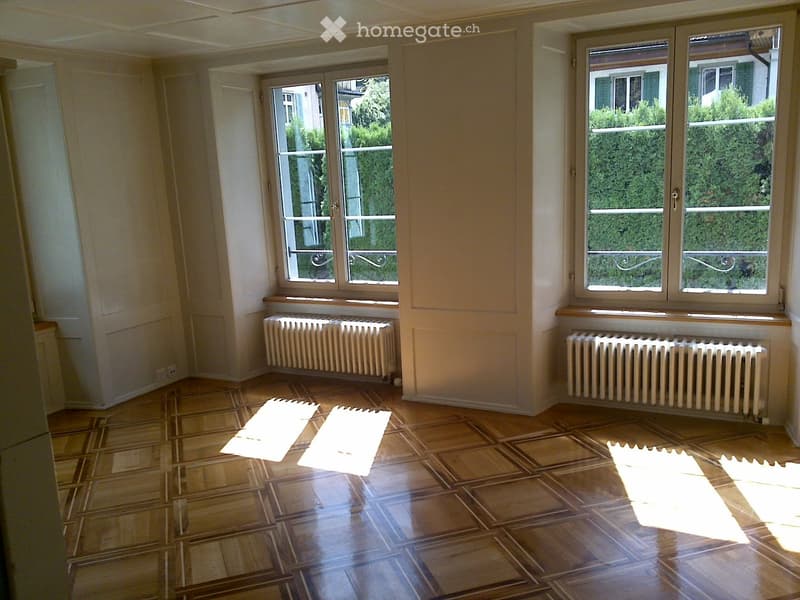 Empty room with wooden flooring, large windows, radiators, and natural light. No furniture or equipment present.
