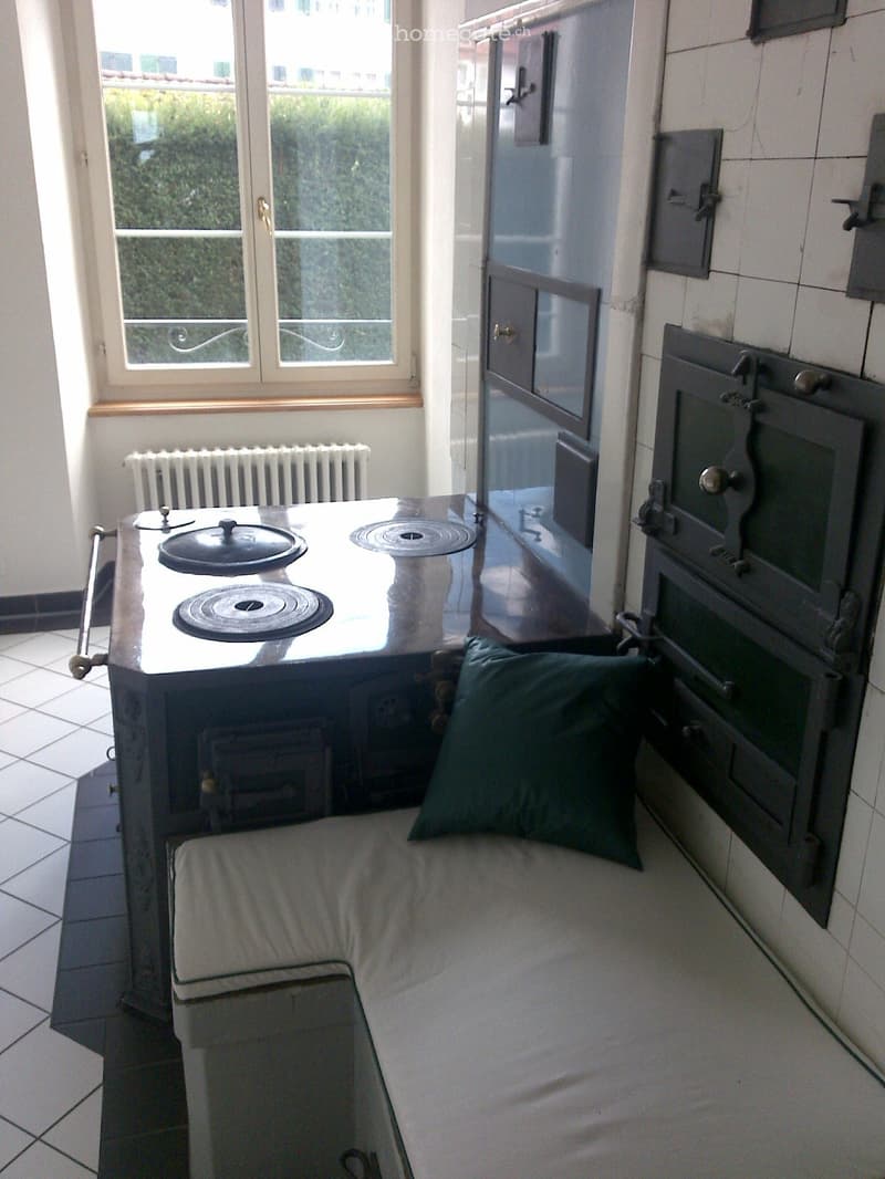 A small, cozy room with a sofa, a table, a radiator, and an oven. The room has tiled flooring and a window with a view of greenery outside.