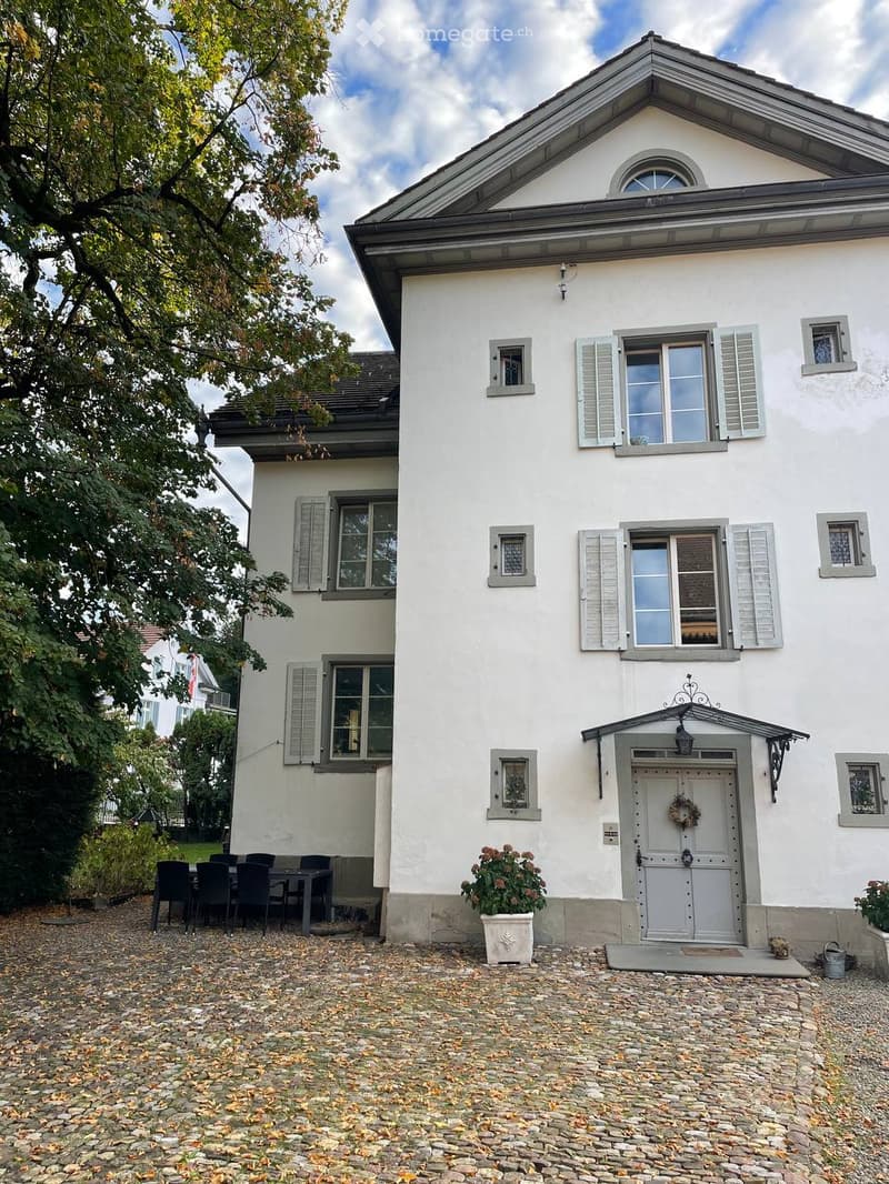 Two-story white house with grey shutters, multiple windows, and a front door adorned with a wreath. A small patio area features a dining set with chairs and a table.