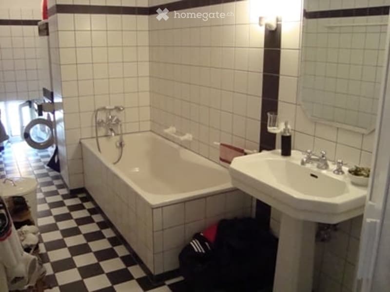 Bathroom with a white tiled wall, black and white checkered floor, a bathtub, a sink, a mirror, and a light fixture on the wall.