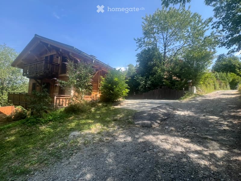 A wooden house with a large balcony, surrounded by trees and a gravel driveway.