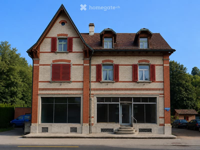 Two-story brick building with red shutters, large windows, and a triangular roof. The ground floor has large glass windows and a central entrance with steps.
