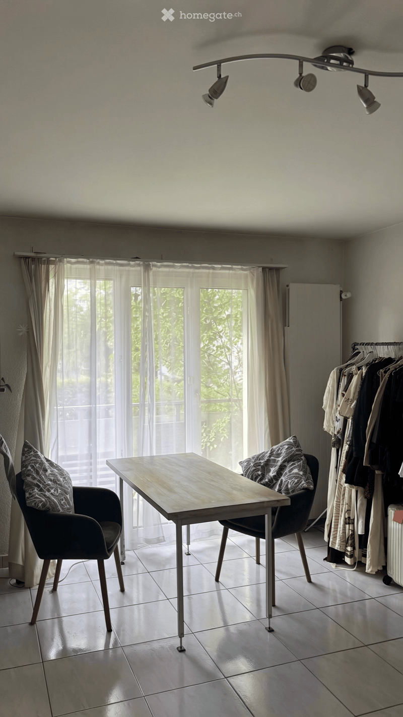 Bright room with tiled floor, large window with sheer curtains, modern table and chairs, clothing rack with hanging clothes.