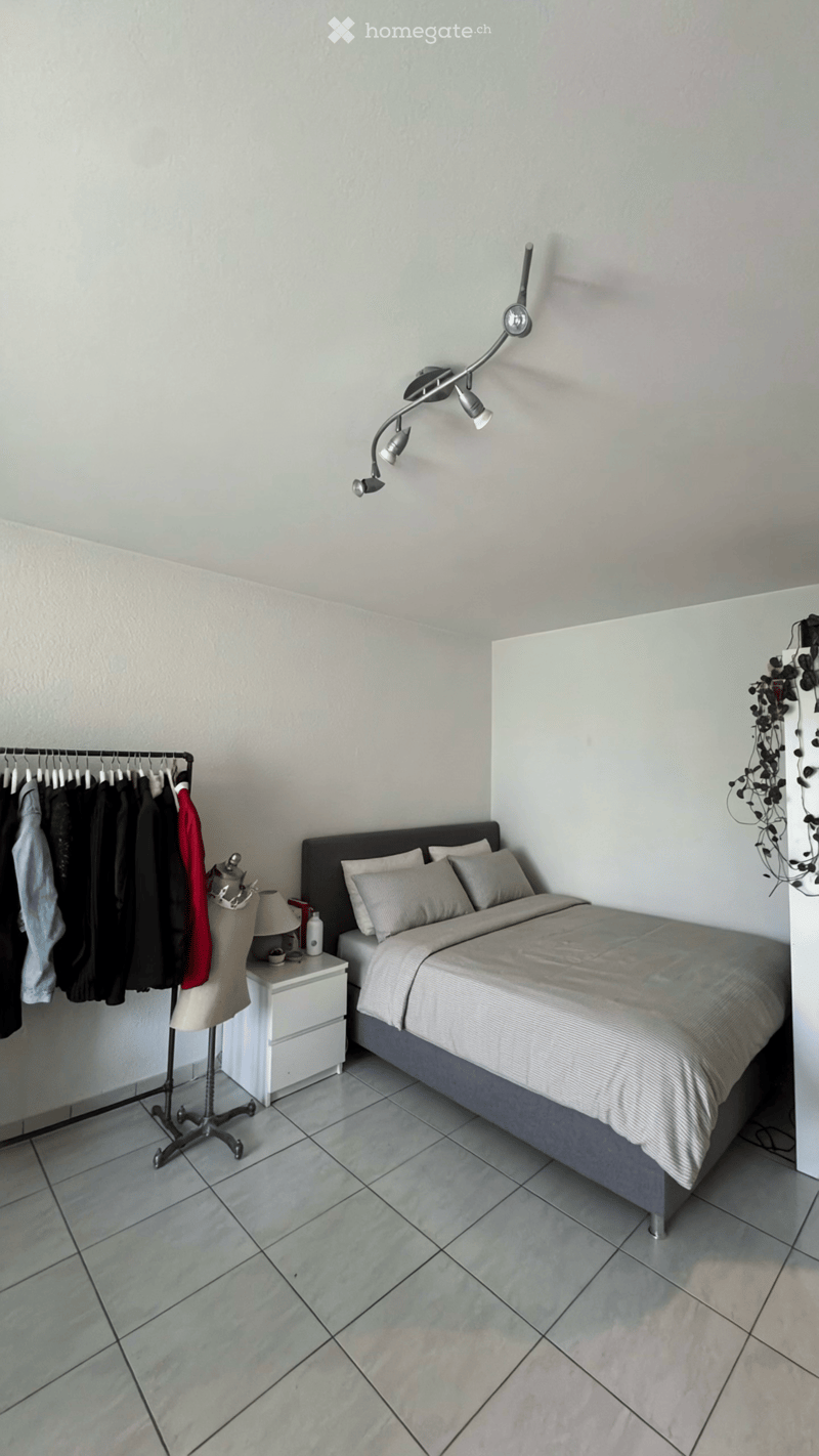 Modern bedroom with a large bed, nightstand, lamp, wardrobe, coat rack, and mannequin. The room features tiled flooring and a sleek, contemporary design.