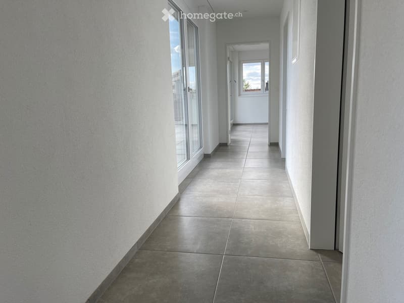 Empty hallway with tiled floor, white walls, and large windows on one side, leading to an open door at the end.