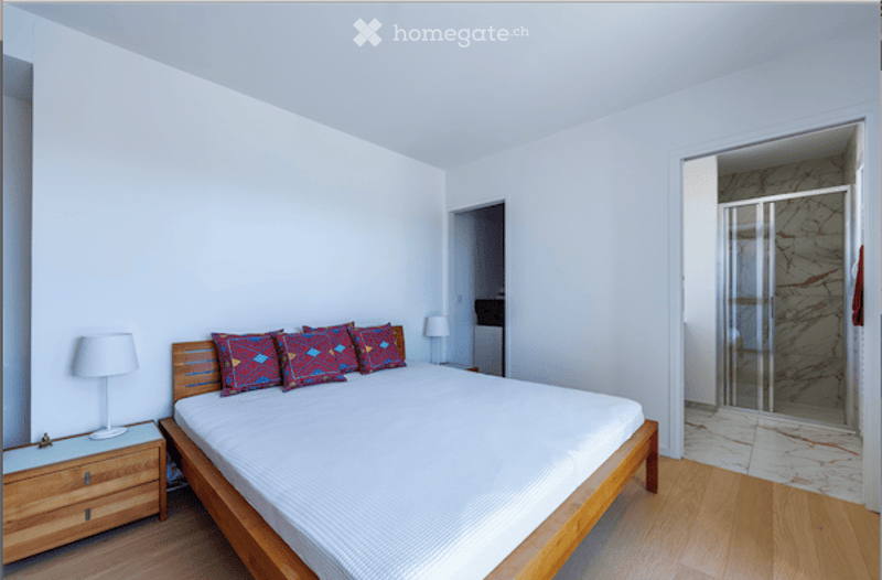 Bright bedroom with a wooden bed frame, white bedding, colorful pillows, a wooden nightstand, and a lamp. A glass-enclosed shower is visible in the adjacent bathroom area.
