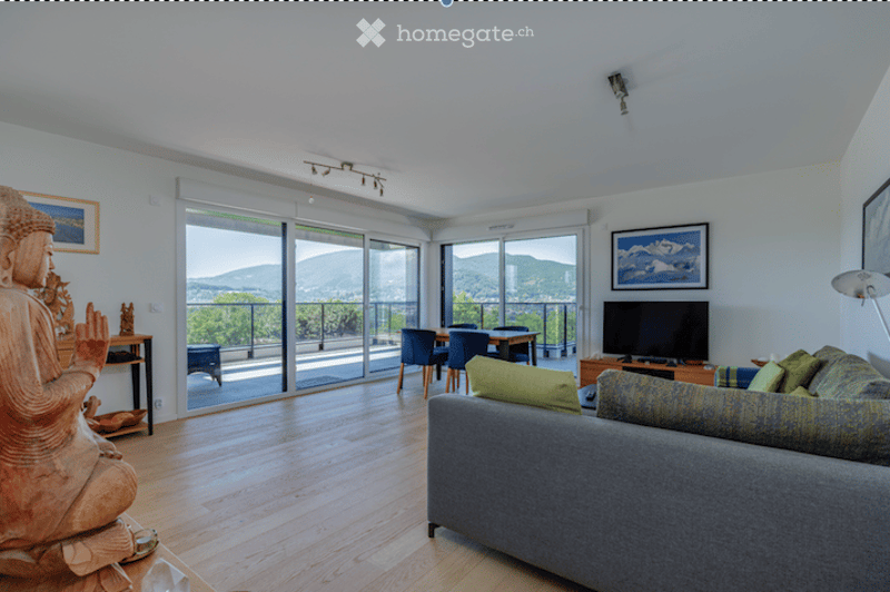 Bright living room with large sliding glass doors opening to a balcony, offering a scenic mountain view. Features a grey sectional sofa, coffee table, dining area with chairs and table, television, floor lamp, and a decorative statue.
