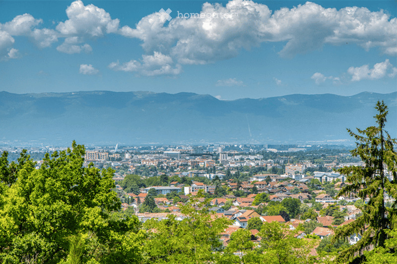 Panoramic view of a cityscape with a mix of residential and commercial buildings, surrounded by green trees and distant mountains under a partly cloudy sky.