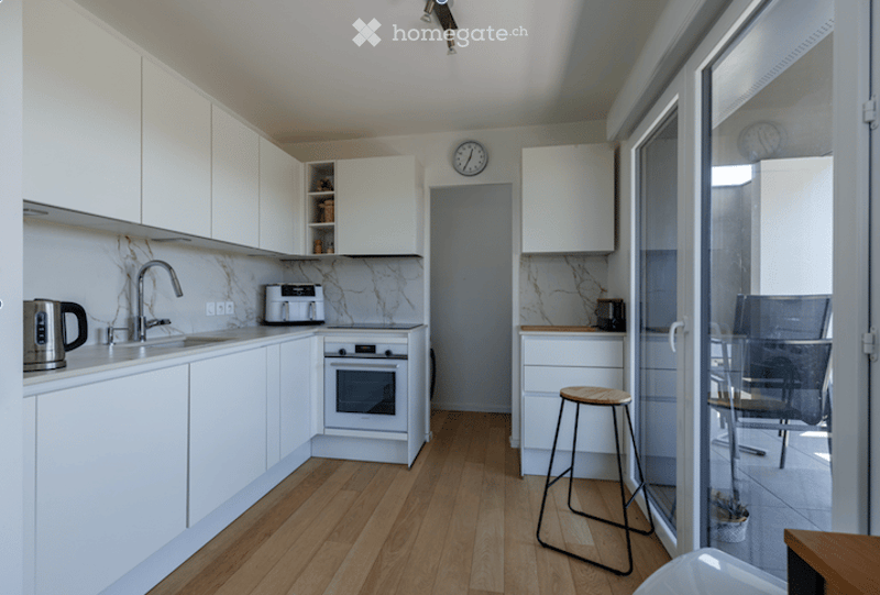 Modern kitchen with white cabinets, marble countertops, wooden flooring, and a small seating area with a stool and chair near a large glass door leading to a balcony.