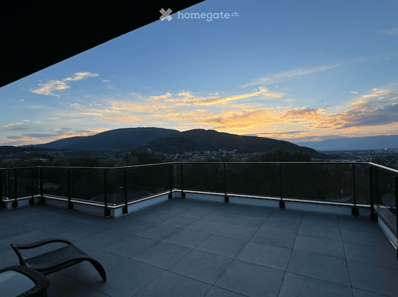 Balcony with a panoramic view of a cityscape and mountains at sunset, featuring a tiled floor and glass railing.