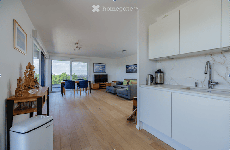 Open-plan living and dining area with wooden flooring, large windows offering a view, a grey sofa, coffee table, TV stand, dining table with chairs, and a modern kitchen area with white cabinets and stainless steel appliances.