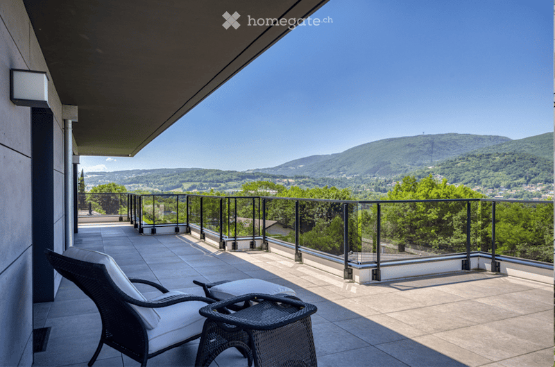 Balcony with modern glass railing, tiled floor, and scenic mountain view. Features a wicker lounge chair and small table.