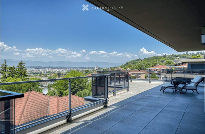 Spacious balcony with modern glass railing, tiled floor, and outdoor furniture including two lounge chairs and a small table, overlooking a scenic cityscape with mountains in the background.
