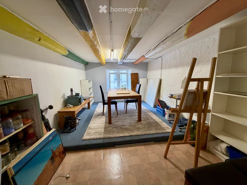 A furnished room with a wooden table, chairs, shelves, a stool, an easel, and a painting on the wall.