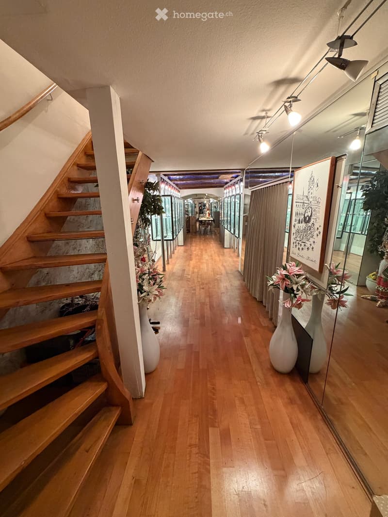 Spacious hallway with wooden flooring, wooden staircase on the left, large white vases with flowers, and framed artwork on the right wall.