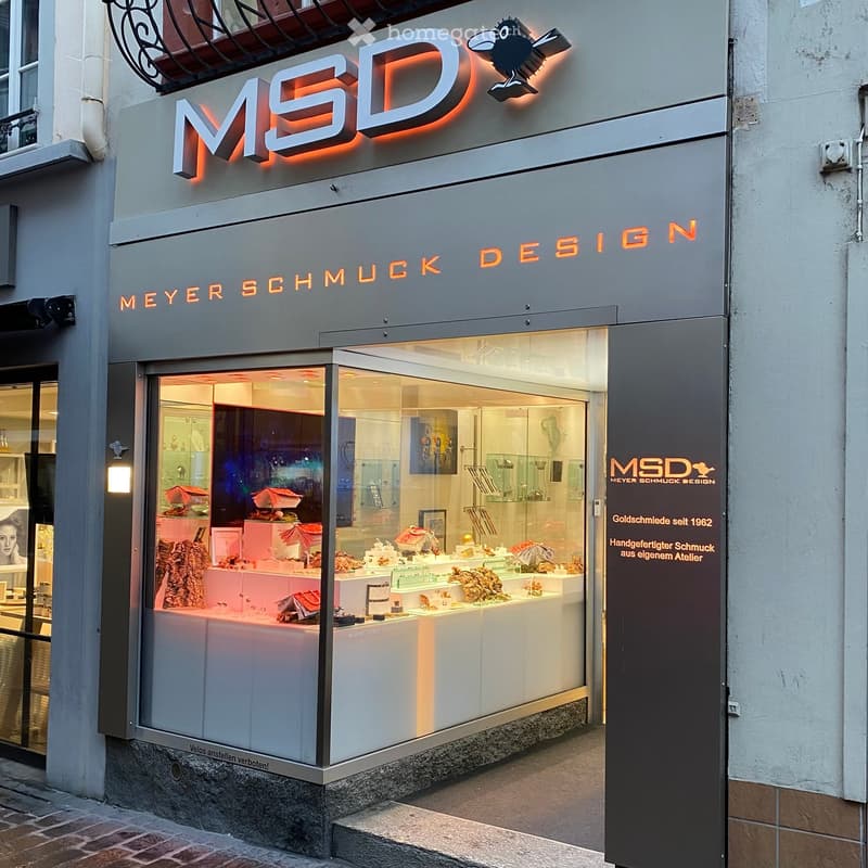 Storefront of Meyer Schmuck Design with illuminated signage, glass display window showcasing jewelry and decorative items, and a modern urban setting.