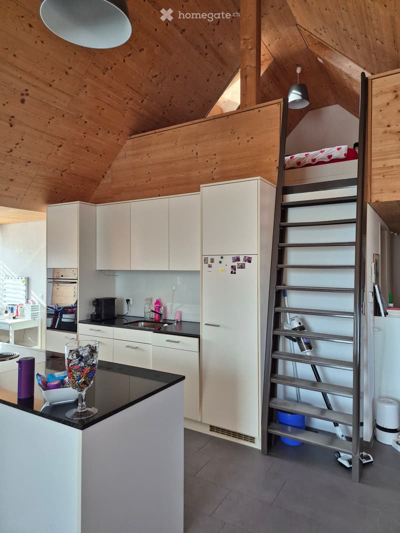 Modern kitchen with white cabinets, black countertops, and a staircase leading to a lofted bedroom area with a bed and lamp.