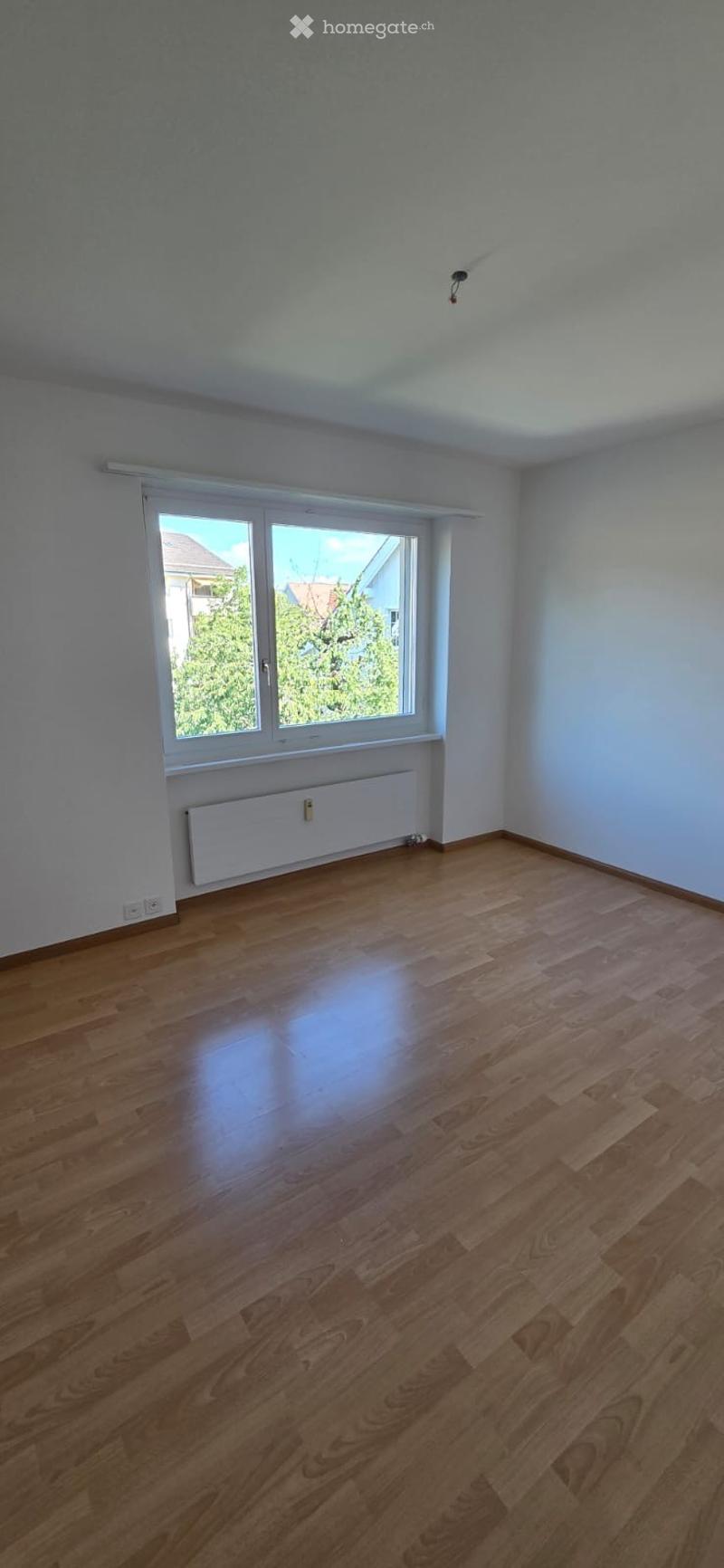 Empty room with wooden flooring, white walls, and a large window with white frame and blinds. A white radiator is placed below the window.