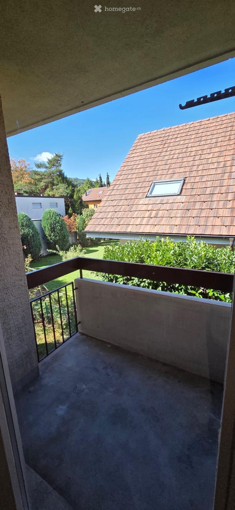 Balcony with a view of a residential area, featuring a wooden railing, concrete floor, and surrounding greenery.