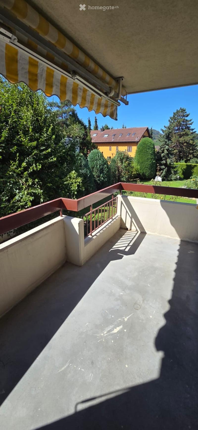 Balcony with a striped awning, metal railing, and a view of a green landscape and a yellow house in the background.