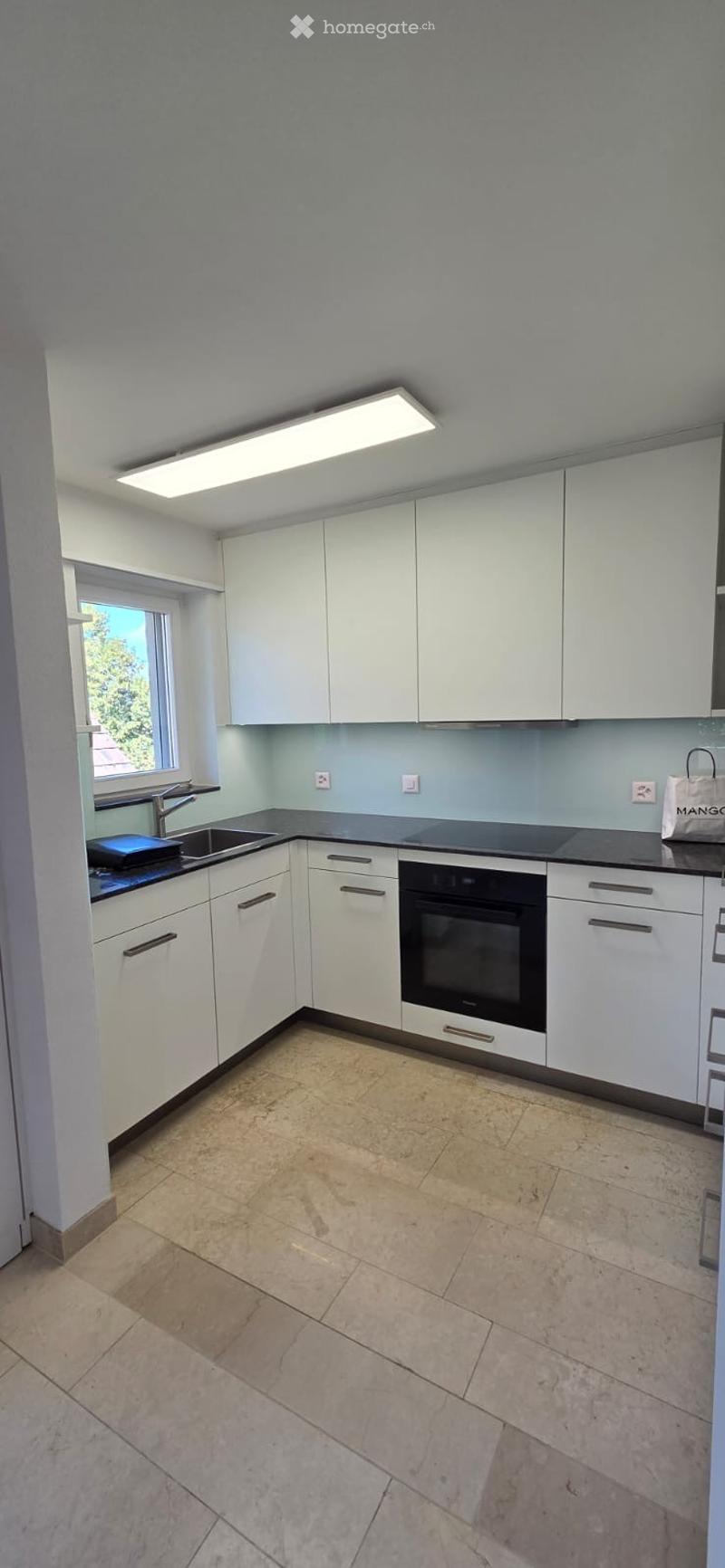 Modern kitchen with white cabinets, a black countertop, a sink, and an oven. The space features tiled flooring and a large window letting in natural light.