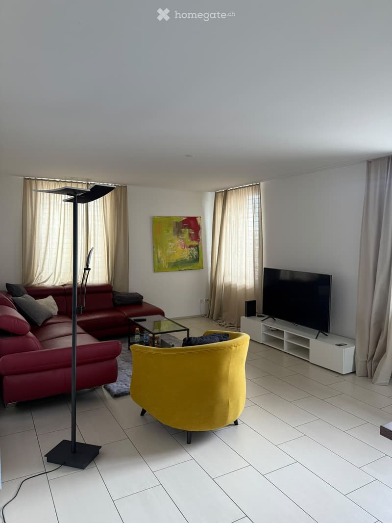 Modern living room with a red sectional sofa, yellow armchair, glass coffee table, flat-screen TV on a white stand, and a floor lamp. The room features large windows with beige curtains and a colorful abstract painting on the wall.