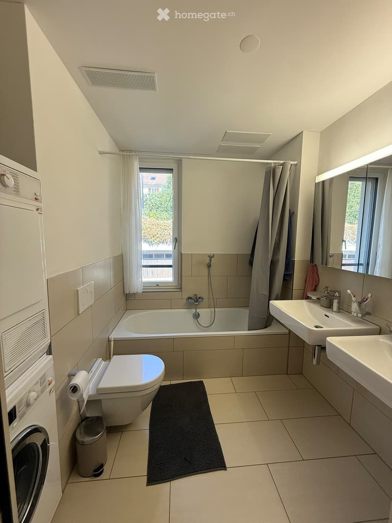 Modern bathroom with a bathtub, toilet, double sink, mirror, washing machine, and a window with a curtain. Includes a black bathroom rug and a toothbrush holder on the sink.