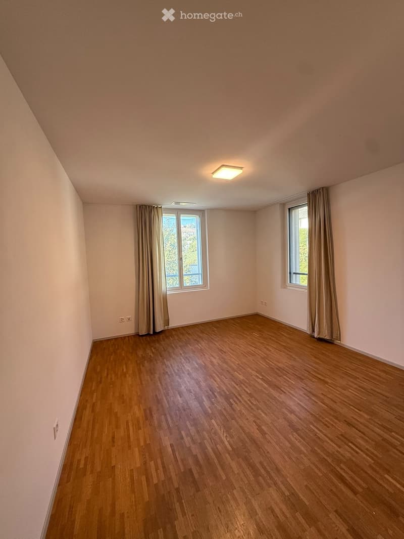 Empty room with wooden flooring, light-colored walls, and two windows with curtains. The room has a ceiling light fixture and electrical outlets on the walls.