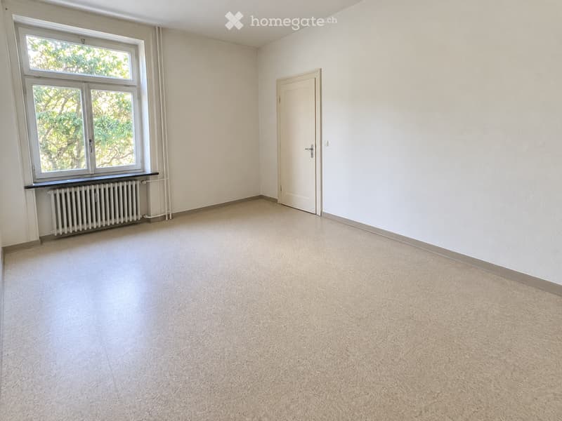 Empty room with light-colored walls, carpeted floor, a window with white frames, a radiator, and a closed door.