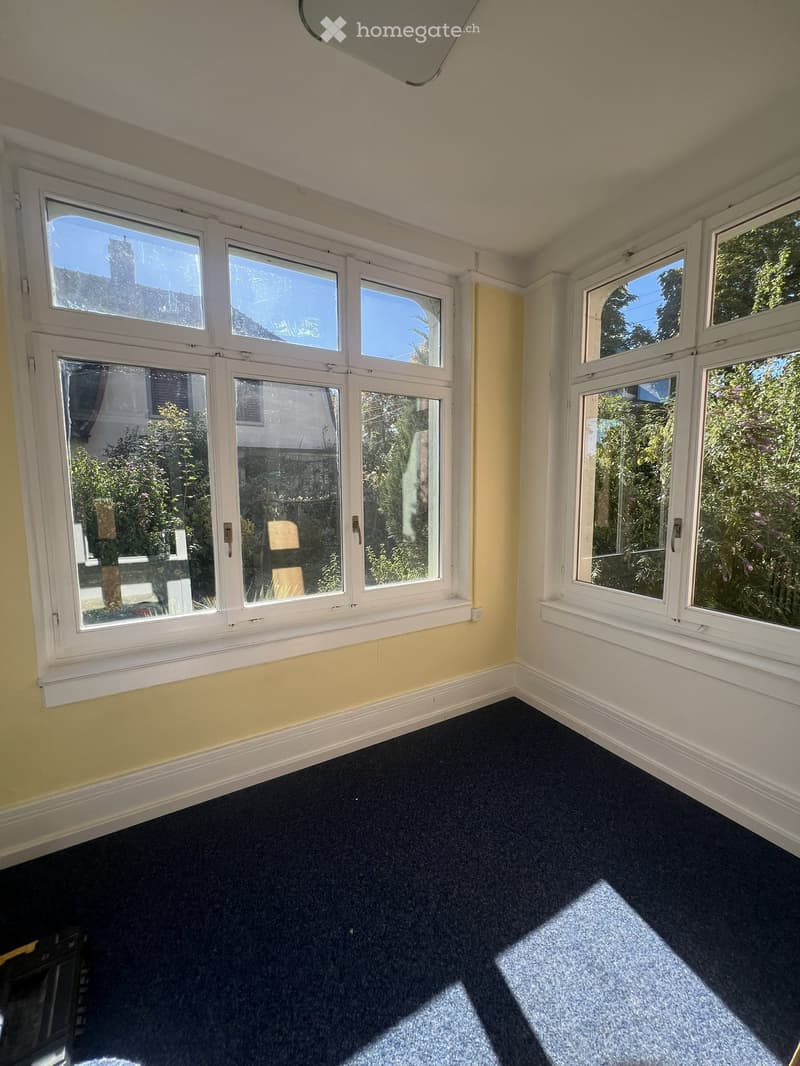 Empty room with dark carpet, white baseboards, and large windows with white frames, allowing natural light to enter.