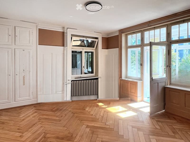 Empty room with wooden herringbone flooring, white walls, large windows, and a radiator. No furniture or personal items present.