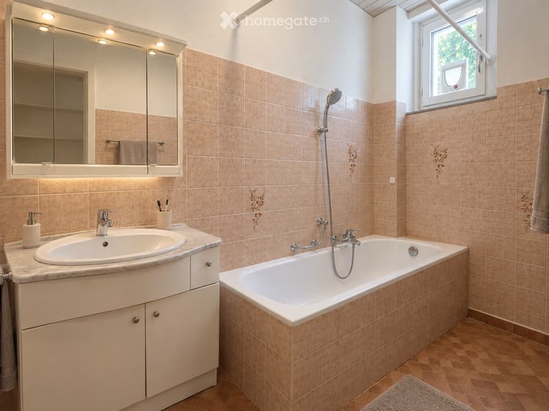Bathroom with beige tiled walls, a white bathtub, a sink with a modern faucet, a large mirror with built-in lighting, and a window with blinds.