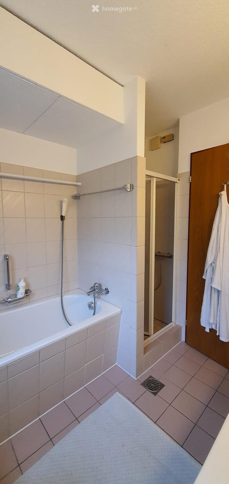 Bathroom with a bathtub, shower area, towel hanging on the door, and a small bottle on the edge of the bathtub.