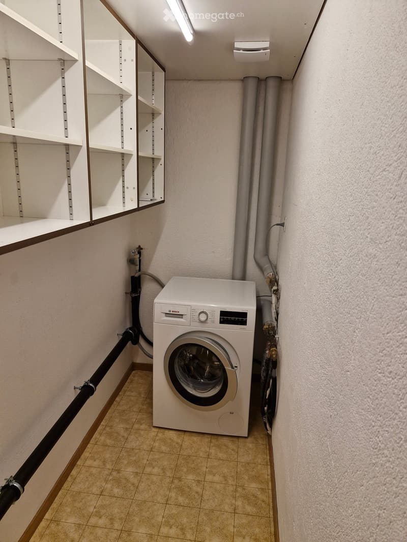 A small, clean utility room with a white Bosch washing machine, tiled floor, and built-in shelving unit.