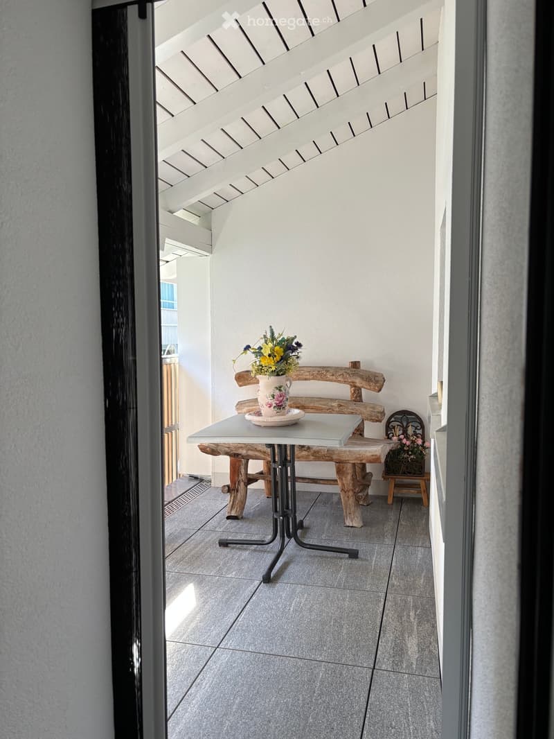 Balcony with a grey tiled floor, a white table with a vase of yellow and pink flowers, a wooden bench, and a small wooden stool with additional flowers.
