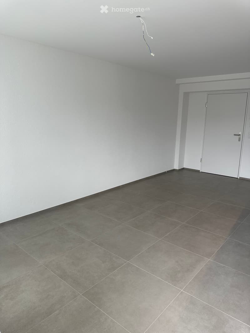 Empty room with grey tiled floor and white walls, featuring a white door and electrical outlet visible on the wall.