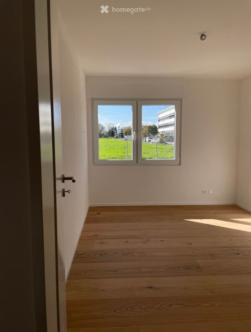Empty room with light wooden flooring, white walls, and a large window offering a view of a green field and a building outside.