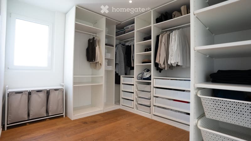 A well-organized walk-in closet with white built-in wardrobes, multiple shelves, drawers, and hanging space for clothes. The floor is wooden, and there is a small laundry basket unit on the left side.
