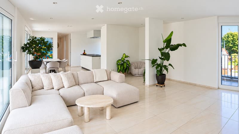 Modern living room with a large sectional sofa, coffee table, dining area with chairs and table, and potted plants. Large windows offer a view of greenery outside.