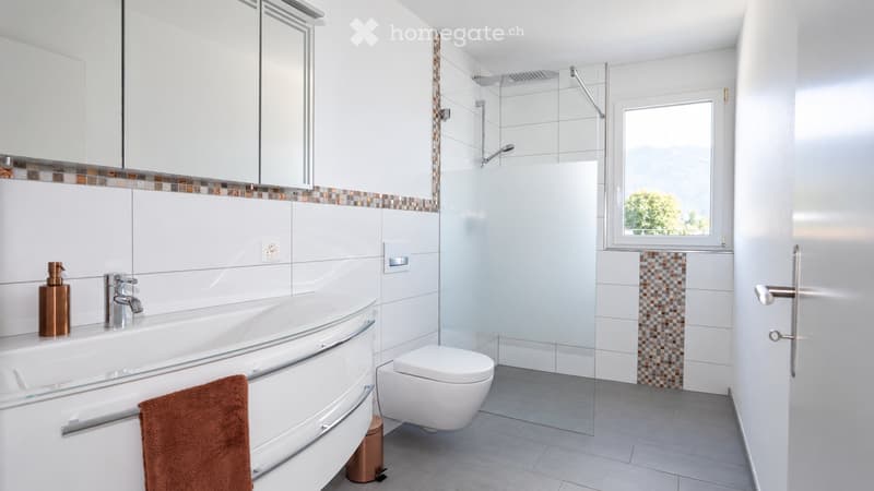 Modern bathroom with white tiles, a bathtub, toilet, and a glass shower enclosure. Features a large window with a view, a towel on the side, and a soap dispenser on the sink.